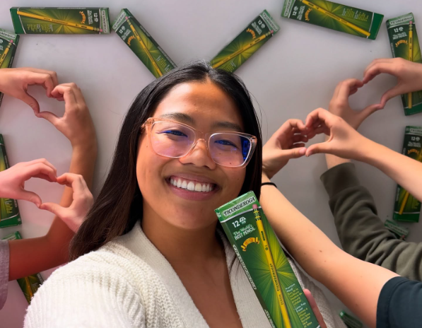 Smiling woman holds product surrounded by friends making heart shapes.