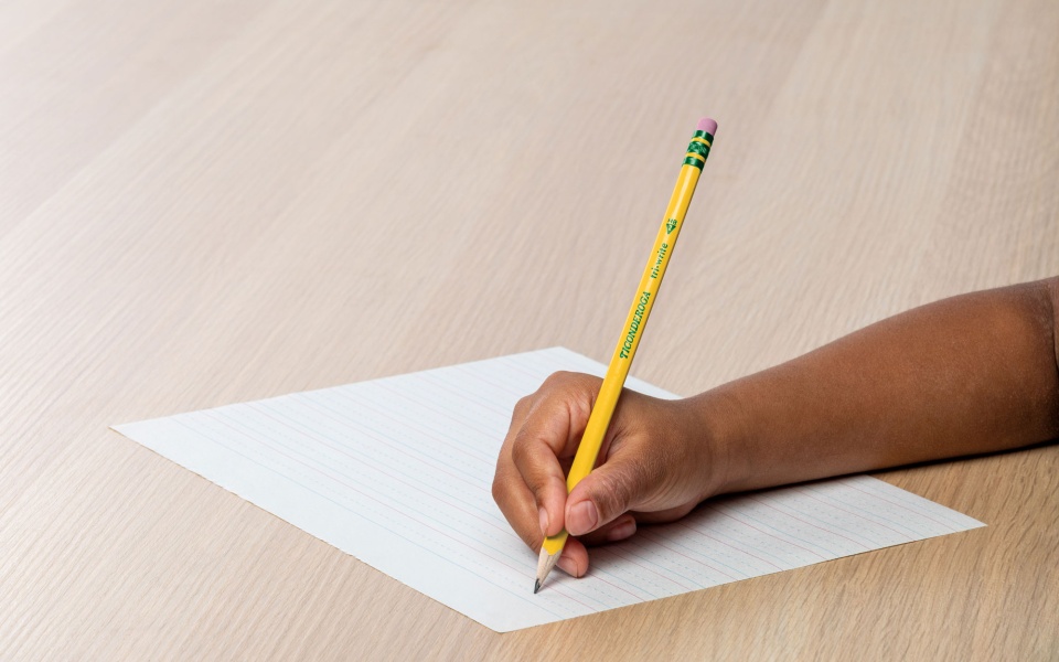 Child writing with a Ticonderoga pencil on lined paper.